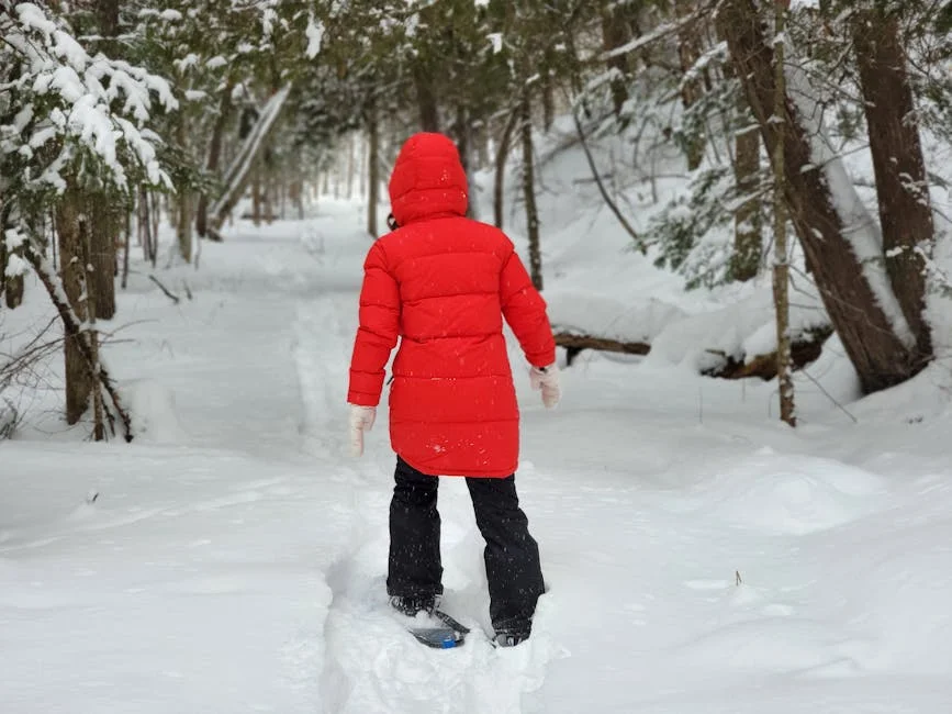 Person in red winter coat snowshoeing through a snowy forest trail surrounded by snow-covered trees