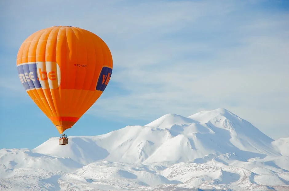 Orange hot air balloon floating above snow-covered mountain peaks under a clear winter sky